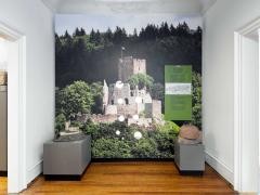 An einer Wand ist eine Fototapete mit einem Bild der Kastelburg. Davor stehen verschiedene Ausstellungsstücke.